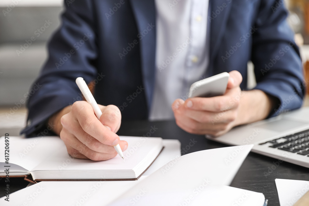 Young businessman with mobile phone writing at table in office, closeup