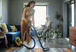 © AnnaStills - Cute teenage girl in casualwear cleaning floor of living room with vacuum cleaner against her grandmother with disability sitting in wheelchair