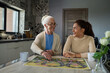 © AnnaStills - Cheerful teenage girl looking at smiling grandmother while sitting by table in the kitchen in front of one another and playing board game at leisure