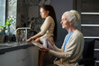 © AnnaStills - Grey haired grandmother with disability wiping clean plates with dry towel while sitting in wheelchair against her granddaughter washing dishes