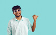 Young cheerful unshaven Indian man in sunglasses smiling pointing with thumb at space for your inscription or advertising dressed in white shirt stands on turquoise studio background