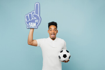 Young surpirsed man fan in t-shirt foam 1 fan glove finger up point overhead cheer up support football sport team hold soccer ball watch tv live stream isolated on plain pastel blue color background