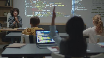 over-the-shoulder full shot of caucasian female teacher sitting at desk in classroom asking student 