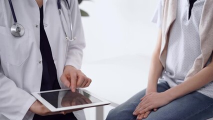 Wall Mural - Doctor and kid patient sitting opposite each other in clinic, close up. Unknown female physician using tablet computer near little boy. Medicine concept