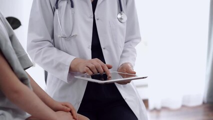 Wall Mural - Doctor and kid patient sitting opposite each other in clinic, close up. Unknown female physician using tablet computer near little girl. Medicine concept