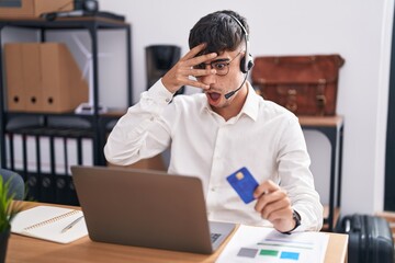 Wall Mural - Young hispanic man working using computer laptop holding credit card peeking in shock covering face and eyes with hand, looking through fingers with embarrassed expression.