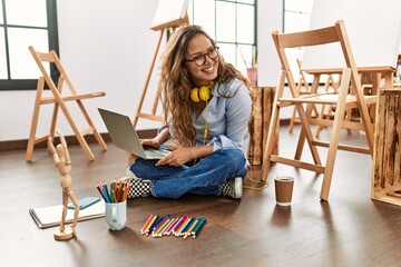 Wall Mural - Young beautiful hispanic woman artist using laptop sitting on floor at art studio