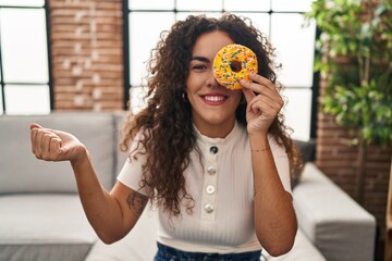 Sticker - Young hispanic woman holding tasty colorful doughnut  eye screaming proud, celebrating victory and success very excited with raised arm