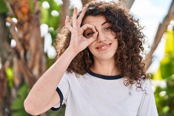 Sticker - Hispanic woman with curly hair standing outdoors smiling happy doing ok sign with hand on eye looking through fingers