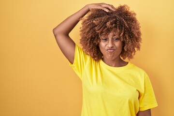 Sticker - Young hispanic woman with curly hair standing over yellow background confuse and wonder about question. uncertain with doubt, thinking with hand on head. pensive concept.