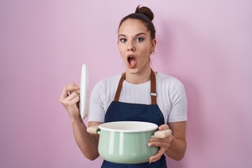 Poster - Young hispanic girl wearing apron holding cooking pot afraid and shocked with surprise and amazed expression, fear and excited face.