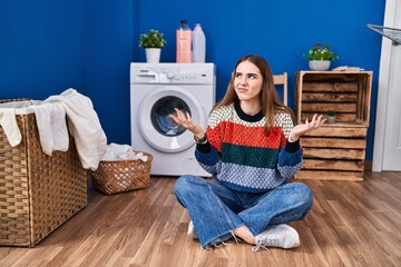 Poster - Young hispanic girl doing laundry clueless and confused expression with arms and hands raised. doubt concept.