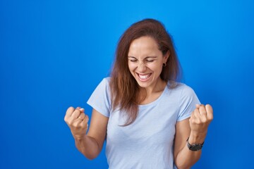 Poster - Brunette woman standing over blue background very happy and excited doing winner gesture with arms raised, smiling and screaming for success. celebration concept.