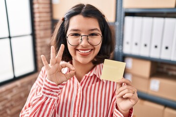 Sticker - Young hispanic woman holding paper reminder at the office doing ok sign with fingers, smiling friendly gesturing excellent symbol