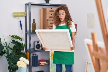 Poster - Young caucasian woman looking at canvas afraid and shocked with surprise and amazed expression, fear and excited face.