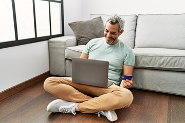 Wall Mural - Middle age grey-haired man using laptop and credit card sitting on floor at home