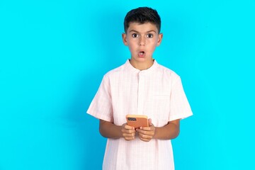 Wall Mural - Shocked Little hispanic boy wearing white shirt  opens mouth hold phone reading advert unbelievable big shopping prices