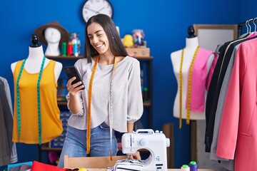 Wall Mural - Young beautiful hispanic woman tailor smiling confident using smartphone at clothing factory