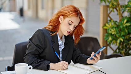 Young redhead woman business worker using smartphone taking notes at coffee shop terrace