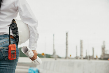 Wall Mural - Young woman engineer or construction worker holding safety hat (helmet), portable radio, and blueprint. Safety Officer inspect in front of oil refinery plant.