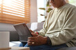 © saltdium - The Close up image features of middle aged Asian man wearing a long-sleeved sweater sitting on a sofa and using digital tablet at home.