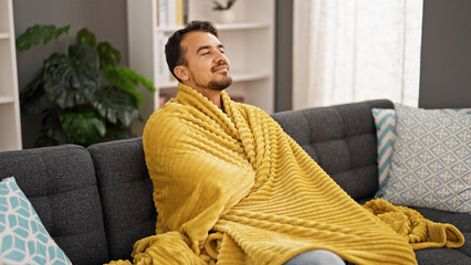 Young hispanic man feeling cold covered with blanket at home