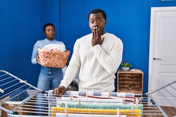 Wall Mural - Young african american couple hanging clothes at clothesline covering mouth with hand, shocked and afraid for mistake. surprised expression