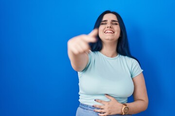 Wall Mural - Young modern girl with blue hair standing over blue background laughing at you, pointing finger to the camera with hand over body, shame expression