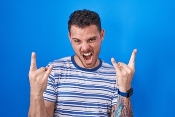 Poster - Young hispanic man standing over blue background shouting with crazy expression doing rock symbol with hands up. music star. heavy music concept.