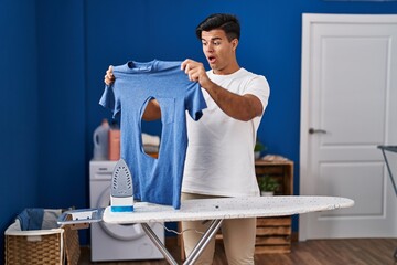 Wall Mural - Hispanic man ironing holding burned iron shirt at laundry room in shock face, looking skeptical and sarcastic, surprised with open mouth