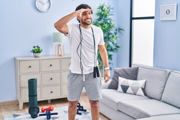 Poster - Handsome latin man wearing sportswear at home very happy and smiling looking far away with hand over head. searching concept.