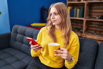Wall Mural - Young blonde woman using smartphone drinking coffee at home