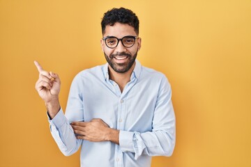 Canvas Print - Hispanic man with beard standing over yellow background with a big smile on face, pointing with hand and finger to the side looking at the camera.