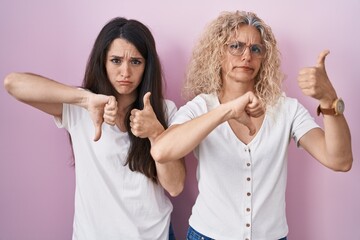 Canvas Print - Mother and daughter standing together over pink background doing thumbs up and down, disagreement and agreement expression. crazy conflict