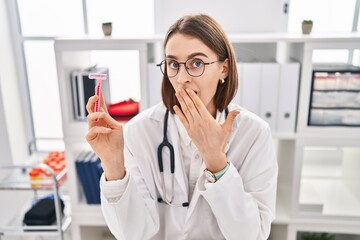 Poster - Young caucasian doctor woman holding razor covering mouth with hand, shocked and afraid for mistake. surprised expression