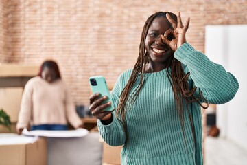 Canvas Print - African woman using smartphone at new home smiling happy doing ok sign with hand on eye looking through fingers