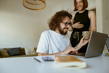 Wall Mural - Young smiling man working on laptop from home while girlfriend is brought him coffee