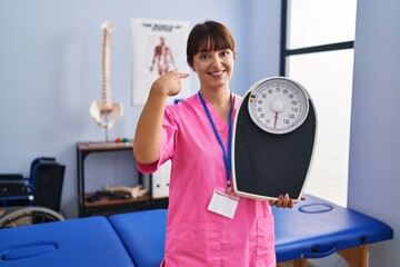 Poster - Young brunette woman as nutritionist holding weighing machine pointing finger to one self smiling happy and proud