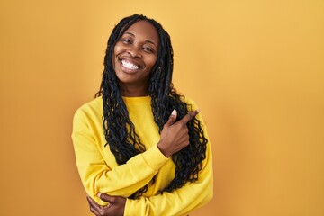 Sticker - African woman standing over yellow background with a big smile on face, pointing with hand and finger to the side looking at the camera.