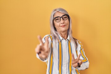 Wall Mural - Middle age woman with grey hair standing over yellow background wearing glasses smiling looking to the camera showing fingers doing victory sign. number two.