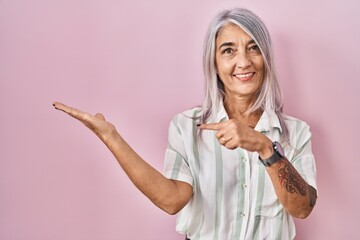 Wall Mural - Middle age woman with grey hair standing over pink background amazed and smiling to the camera while presenting with hand and pointing with finger.