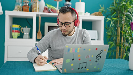 Poster - Hispanic man wearing headphones using laptop writing on notebook at dinning room