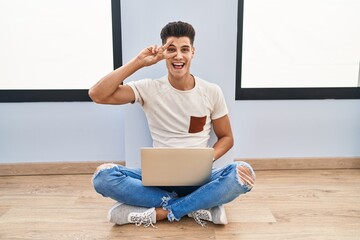 Sticker - Young hispanic man using laptop at home doing peace symbol with fingers over face, smiling cheerful showing victory