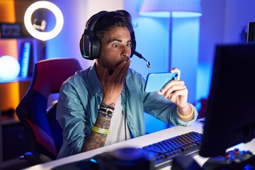 Poster - Young hispanic man with tattoos playing video games with smartphone covering mouth with hand, shocked and afraid for mistake. surprised expression