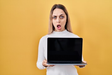 Wall Mural - Young blonde woman holding laptop in shock face, looking skeptical and sarcastic, surprised with open mouth