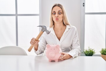 Wall Mural - Young blonde woman holding piggy bank and hammer making fish face with mouth and squinting eyes, crazy and comical.