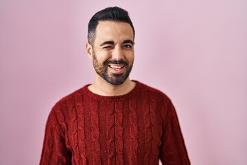 Poster - Young hispanic man with beard wearing casual sweater over pink background winking looking at the camera with sexy expression, cheerful and happy face.