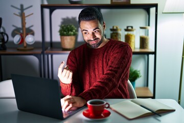 Canvas Print - Young hispanic man with beard using computer laptop at night at home beckoning come here gesture with hand inviting welcoming happy and smiling