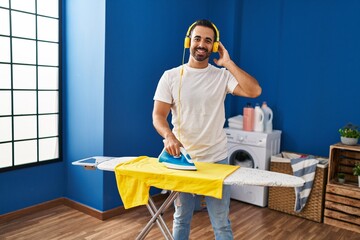 Wall Mural - Young hispanic man listening to music ironing clothes at laundry room