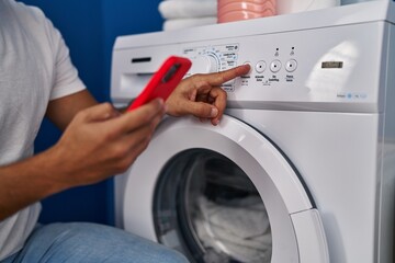 Wall Mural - Young hispanic man turning on washing machine using smartphone at laundry room